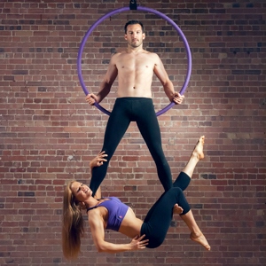 Cerchio aereo viola X-Pole davanti a una parete di mattoni con una ballerina appesa alle gambe di un ballerino